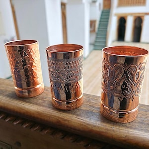 May include: Three copper-coloured drinking glasses with embossed patterns. The glasses are cylindrical, featuring geometric and floral designs. They are displayed on a wooden surface, highlighting their reflective, metallic finish. Each glass has a unique design.