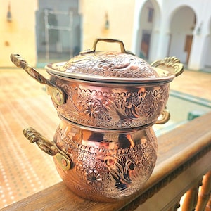 Pode incluir: Conjunto de panelas de cobre de duas peças com padrões florais e de folhas ornamentados. O conjunto inclui uma panela com tampa e uma panela menor empilhada por baixo. Ambas as panelas têm pegas decorativas.