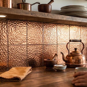 May include: A kitchen interior with copper elements. The backsplash is made of hammered copper tiles. A copper kettle, pots, and other kitchenware are arranged on a wooden countertop and shelf. The style is rustic and inviting.