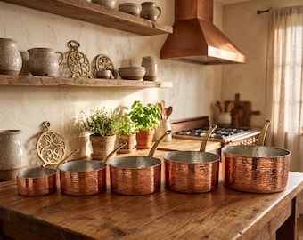 May include: A set of five copper saucepans with brass handles arranged on a wooden surface. The hammered copper pots vary in size, with the largest measuring approximately 10 inches in diameter. The kitchen setting includes shelves with pottery and plants.