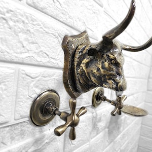 May include: A bronze-toned wall-mounted faucet with a bull head design. The faucet features a decorative bull head plaque and cross-shaped handles. The fixture is mounted on a white brick wall, creating a rustic aesthetic.