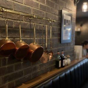 May include: Copper pans hanging on a brass rail against a brick wall. The pans have brass handles and are different sizes. The wall is made of dark brown bricks. The scene is in a restaurant.