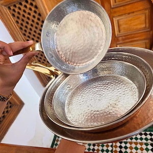 Op de afbeelding: Een set gehamerde metalen pannen met messing handvatten. De pannen, in verschillende maten, hebben een getextureerd oppervlak. De pannen zijn gestapeld, met één omhooggehouden. De achtergrond bevat houten architecturale details.