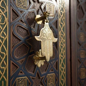 May include: Close-up of a wooden door with intricate geometric carvings and a gold-colored Hamsa hand-shaped door knocker. The door features dark brown, blue, and gold accents, with Arabic calligraphy in square panels. The design is ornate and detailed.