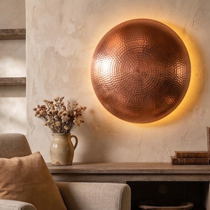 May include: A large, hammered copper wall sconce with a warm, ambient glow. The round fixture is mounted on a textured wall, creating a focal point in a cozy living space. The sconce is approximately 30 inches in diameter.