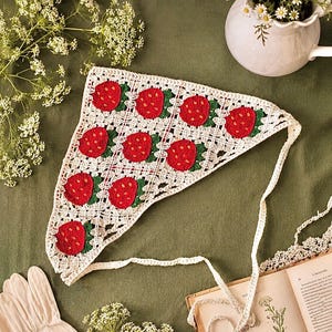 May include: A handmade, white crochet headscarf featuring a repeating pattern of red strawberries with green leaves. The headscarf has a triangular shape and is tied with a long, white crochet string. The background is a muted green.