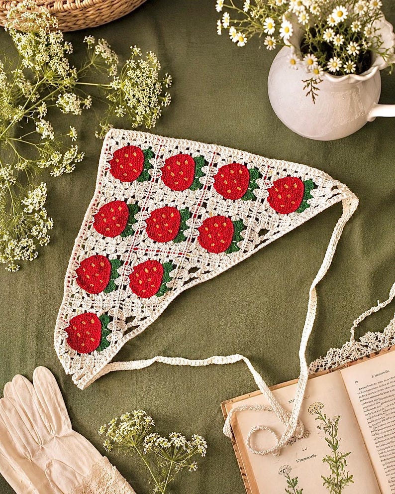 May include: A handmade, white crochet headscarf featuring a repeating pattern of red strawberries with green leaves. The headscarf has a triangular shape and is tied with a long, white crochet string. The background is a muted green.