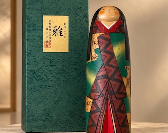 May include: A cylindrical wooden Kokeshi doll with a smooth, rounded head and black hair, wearing a garment featuring green, yellow, and maroon geometric patterns. Beside it stands a tall, textured dark green box with a gold label containing Japanese text.