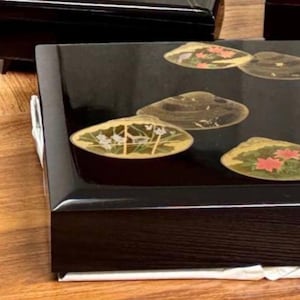 Op de afbeelding: Zwarte gelakte doos met schelpvormige ontwerpen. De doos heeft een glanzende afwerking en is versierd met schelpvormige ontwerpen met bloemen- en plantmotieven. De rechthoekige doos heeft een houten uitstraling.