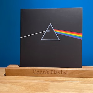 May include: A vinyl record album with a black cover, a white triangle, and a rainbow. It rests on a wooden stand engraved with "Colin's Playlist". The background is a solid blue wall.