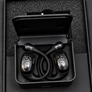 May include: A pair of black wireless earbuds with ear hooks, positioned inside a black rectangular charging case. The earbuds feature a glossy finish on the outer surface and are marked with R and L indicators for right and left orientation.