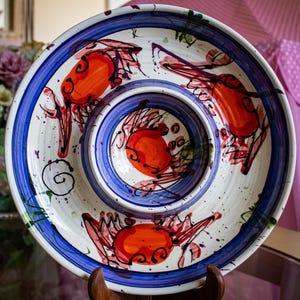 May include: A round ceramic serving dish with a central bowl and a wide rim, featuring hand-painted orange crab illustrations, blue circular bands, and dark purple paint splatters on a cream-coloured background with a glossy finish.