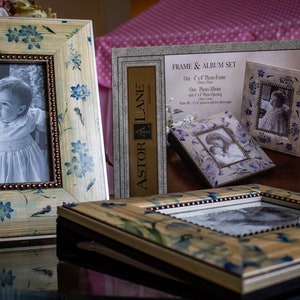 May include: A set of two picture frames with a floral design. The frames are made of wood and have a light brown finish. The frames are decorated with blue flowers and have a beaded border. The frames are displayed on a table with a pink polka dot tablecloth. The packaging for the frames is visible in the background and says "FRAME & ALBUM SET" and "ASTOR LANE".