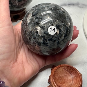 May include: A speckled, dark gray sphere with a white sticker labeled "14" is held in a hand. The sphere has a polished surface with reflective qualities. The item is a decorative stone or crystal ball, likely for display or meditation.