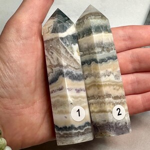 May include: Two banded fluorite crystal towers. The towers are numbered 1 and 2 with a white sticker. The crystals have layers of white, tan, purple, and green. The crystals are held in a hand.