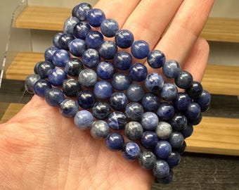 May include: A multi-strand bracelet composed of polished, spherical sodalite beads. The beads exhibit a deep blue color with mottled white and grey inclusions, arranged in a dense, layered formation that covers the palm of a hand.
