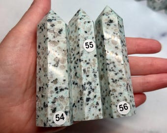 May include: Three K2 stone obelisks with a light blue and speckled pattern. Each obelisk has a pointed top and is labeled with a white sticker with a black number. The obelisks are held in a hand.