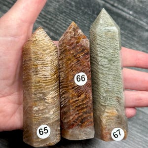 May include: Three polished crystal towers with textured patterns in shades of brown, red, and green. Each tower has a pointed top and a rectangular base. The crystals are labeled with white stickers numbered 65, 66, and 67.