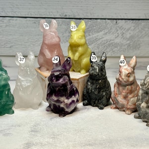 May include: A collection of nine carved stone rabbit figurines in various colors, including green, white, pink, yellow, purple, black, red, and gray. Each rabbit has a unique texture and is numbered with a small white sticker.