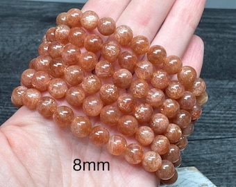 May include: A bracelet of sunstone beads, each 8mm in diameter, is displayed. The beads are a warm, translucent orange with sparkling inclusions. The bracelet is resting on a hand, showcasing the color and texture of the gemstones.