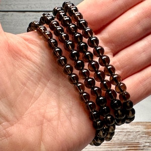 Peut inclure: Quatre rang&eacute;es de perles de pierres pr&eacute;cieuses rondes, brun fonc&eacute;, sont tenues dans une main. Les perles sont enfil&eacute;es pour former un bracelet. Les perles ont une qualit&eacute; translucide et une finition polie. Le bracelet est un accessoire neutre.