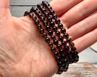 May include: Four strands of dark brown, round gemstone beads are held in a hand. The beads are strung together to form a bracelet. The beads have a translucent quality and a polished finish. The bracelet is a neutral accessory.
