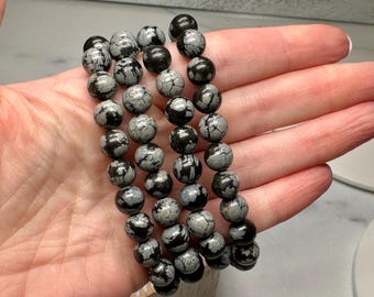 May include: Snowflake obsidian beaded bracelets. The bracelets feature round beads in shades of gray and black. The beads have a speckled pattern. The bracelets are shown in a hand.
