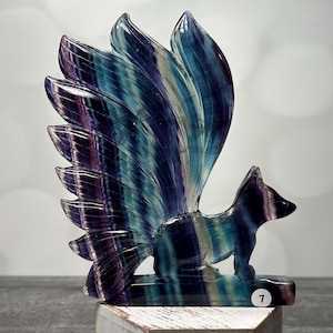 May include: A carved fluorite fox figurine with a large, fanned tail. The fox is predominantly blue, purple, and green, with a polished finish. The figurine is displayed on a wooden surface.