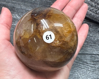 May include: A polished, spherical quartz crystal featuring internal golden-brown inclusions and fractures. A small, circular white sticker with the black number 61 is affixed to the center of the smooth, translucent surface of the stone sphere.
