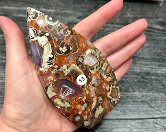 May include: A polished, teardrop-shaped piece of orbicular jasper featuring a complex pattern of brown, rust-orange, cream, and deep purple tones with small crystalline inclusions. A small white circular sticker with the number 13 is attached.