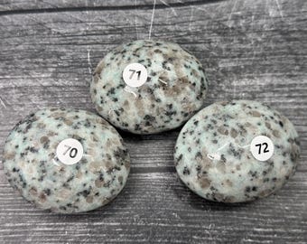 May include: Three polished kyanite stones with a speckled pattern of gray, black, and light green. Each stone has a white sticker with a black number: 70, 71, and 72. The stones are on a gray wooden surface.