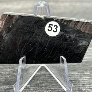 May include: A rectangular, polished stone with a black and brown marbled pattern. A white sticker with the number 53 is affixed to the stone. The stone is displayed on a clear plastic stand.