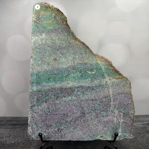 May include: A large, irregularly shaped slab of stone with a mix of green, purple, and red hues. The stone has a rough, textured surface and is displayed on a black metal stand. The colors are layered, creating a unique pattern.