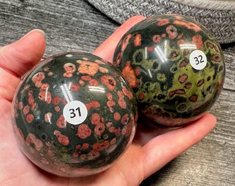 May include: Two polished spherical stones featuring a dark green base with speckled patterns of red and light green. Each sphere has a small white circular sticker applied to the surface, with the number 31 on one and the number 32 on the other.