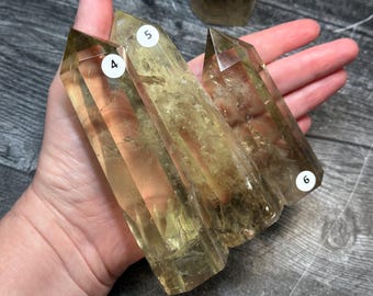 Puede incluir: Tres puntas de cristal de citrino translúcidas de tono amarillo con extremos facetados y puntiagudos. Cada cristal presenta una pequeña pegatina circular blanca etiquetada con los números 4, 5 y 6, indicando opciones de selección.