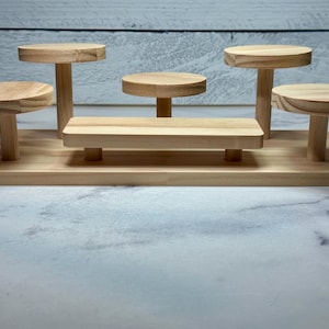 May include: A wooden display stand with multiple platforms of varying heights. The stand features five circular platforms and one rectangular platform, all made of light-colored wood. The platforms are supported by cylindrical legs, creating a tiered effect.