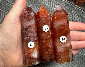 May include: Three polished crystal points with a reddish-brown hue and internal white inclusions. Each point features a hexagonal shape and a pointed top. White circular stickers labeled 12, 13, and 14 are affixed to the front of each crystal.
