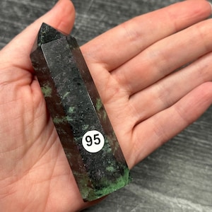May include: A dark green and black crystal point with a faceted design. The crystal has a pointed top and a white sticker with the number 95 on it. The crystal is held in a hand.
