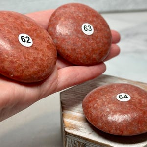 May include: Three smooth, reddish-orange stones with textured surfaces. Two are held in a hand, and one rests on a wooden block. Each stone has a small white sticker with a black number: 62, 63, and 64.