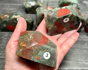 May include: A collection of faceted, natural stone crystals. The crystals are primarily green with red and brown inclusions. Each crystal has a unique pattern and shape. Some crystals have small white stickers with numbers.