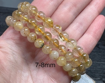 May include: Three golden rutile quartz bracelets, each strung with 7-8mm beads. The beads are translucent with golden inclusions, creating a shimmering effect. The bracelets are displayed on a light background.