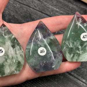 May include: Three teardrop-shaped fluorite crystals in shades of green and purple, each with a white sticker displaying a number. The crystals are held in a hand, showcasing their translucent quality and natural patterns. The crystals are a unique addition to any collection.