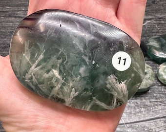 May include: A polished, oval-shaped green and dark purple stone with visible white, feathery, fan-like inclusions throughout the surface. A small, circular white sticker with the black number 11 is affixed to the right side of the stone.