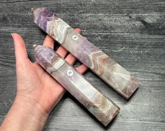 May include: Two amethyst crystal obelisks with a polished finish. The crystals have a mix of purple, white, and brown tones. Each obelisk has a sticker with a number on it. The crystals are held in a hand.