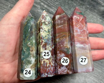 May include: Four polished gemstone obelisks in shades of green, red, and brown are held in a hand. Each obelisk has a pointed top and a rectangular base. White stickers with the numbers 24, 25, 26, and 27 are affixed to the obelisks.