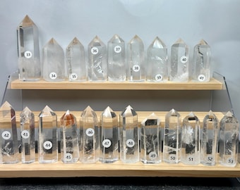 May include: A collection of twenty-one clear quartz crystal points arranged on a two-tiered wooden display stand. Each crystal is upright, faceted, and labeled with a small white circular sticker numbered from 33 to 53 for identification.