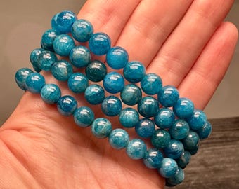 May include: A multi-strand bracelet composed of polished, round blue apatite gemstone beads. The beads exhibit varying shades of vibrant teal and deep ocean blue with natural internal inclusions and a smooth, reflective surface finish.