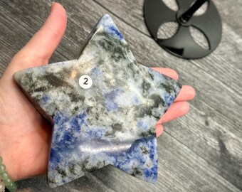 May include: A star-shaped decorative stone with a marbled pattern of blue, gray, and black. The stone is held in a hand and has a small white sticker with the number "2" on it. The background is a wooden surface.