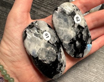 May include: Two polished, oval-shaped moonstone crystals. The stones are primarily white with black and gray marbling, and a hint of blue iridescence. Each stone has a small white sticker with a number on it.
