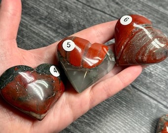 May include: Three heart-shaped stones in shades of red, gray, and black are held in a hand. Each stone has a unique pattern and texture. The stones are numbered with white stickers. The background is a gray wooden surface.